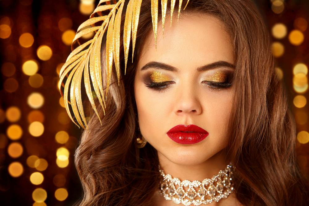 Sexy elegant woman with red lips, glitter eyeshadow in expensive diamond necklace over Christmas lights background | New Year’s Eve Makeup Tutorial | Gold Glitter Cut Crease With Red Lips