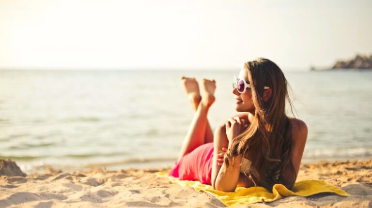 woman smiling while lying on beach sand | How To Tan Safely and Faster For A Glowing Sunkissed Skin | how to tan | Featured